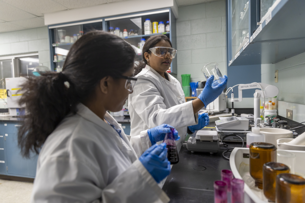 Researchers in white coats and safety glasses work with lab equipment