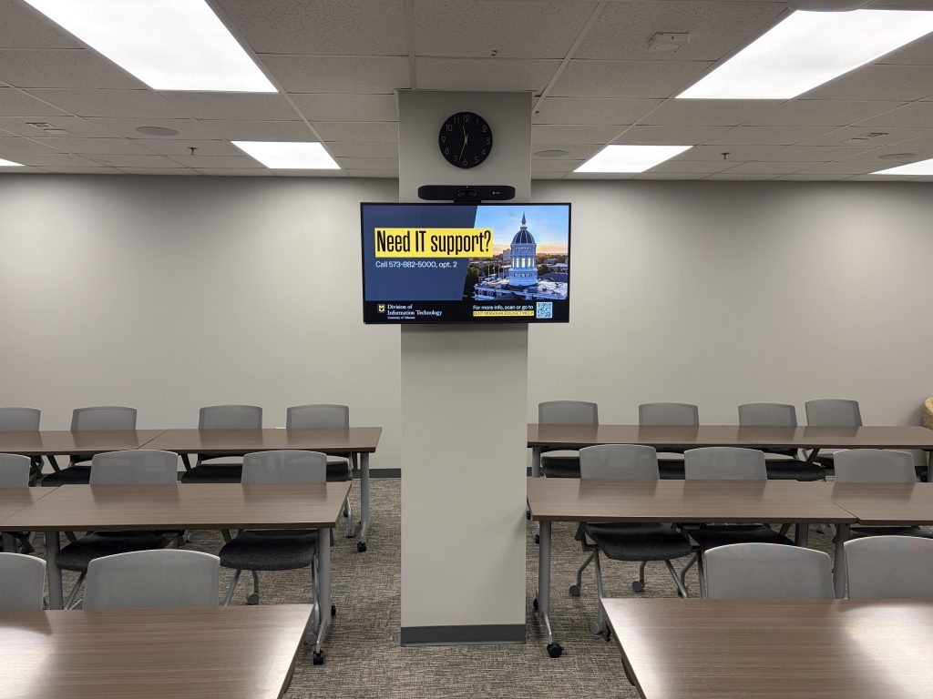 An empty classroom space with chairs and tables from the point of view of a presenter with a pillar and digital screen in the center of the room