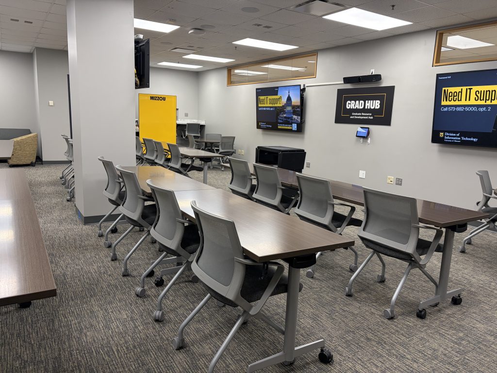 An empty classroom space with chairs and tables facing digital displays with a sign reading Grad Hub in the center